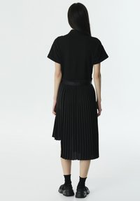 Zwarte geplooide jurk met een kraag bovenaan en korte mouwen, met een strakke taille en een vloeiende rok; gecombineerd met zwarte sneakers.