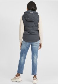 Gilet matelassé gris foncé avec capuche, présentant des sections cousues. Associé à un jean bleu clair et des baskets beiges avec des accents roses.