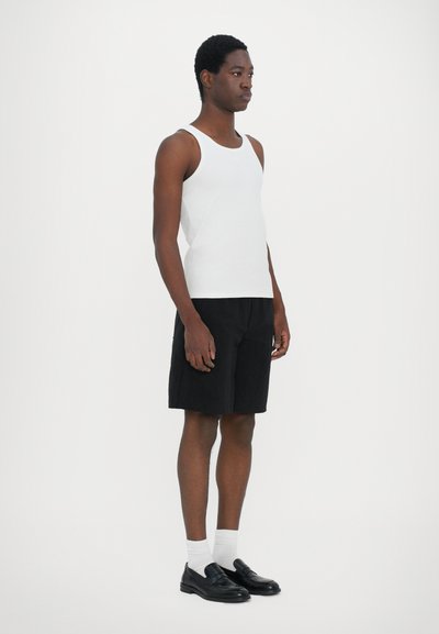 Débardeur blanc à côtes, shorts noirs et chaussures noires à enfiler avec des chaussettes blanches. Le mannequin se tient de profil contre un fond blanc uni.