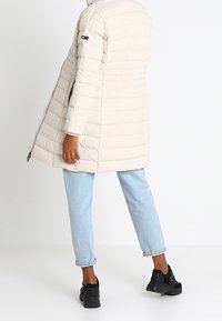 Beige pufferjacka med ett kviltat mönster, långa ärmar, sidofickor med dragkedja och en logotyp på överarmen, kombinerad med ljusblå jeans och svarta sneakers.