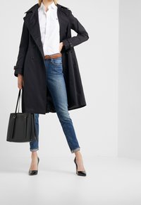 Svart trenchcoat med dubbelknäppning, kombinerad med vit skjorta, blå jeans och svarta högklackade skor. Accessoar: svart handväska.