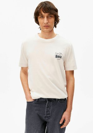 Jonge man met schouderlang bruin haar, gekleed in een wit T-shirt met de tekst "Stap op je fiets" en donkere spijkerbroek, staand tegen een witte achtergrond.