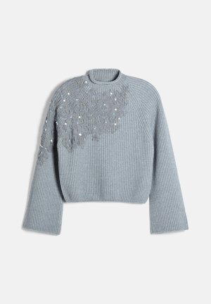 Femme portant un pull côtelé gris avec un détail en dentelle florale, pantalon large en velours côtelé gris, ceinture noire, bottines noires, tenant un sac à main noir.