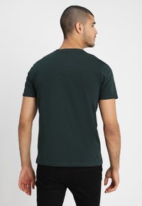 Dunkelgrünes T-Shirt mit kurzen Ärmeln aus weichem Stoff, mit Rundhalsausschnitt und geradem Saum. Getragen mit schwarzen Hosen.