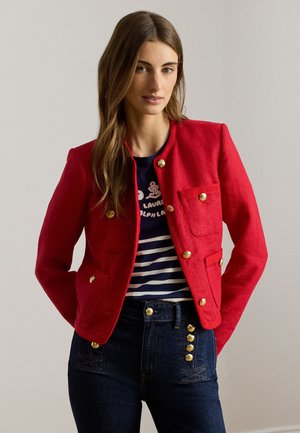 Κόκκινο σακάκι με χρυσά κουμπιά, δύο μπροστινές τσέπες και δομημένο σχέδιο. Φορεμένο πάνω από μπλε-ναυτικό και άσπρο ριγέ πουκάμισο και σκούρο τζιν.