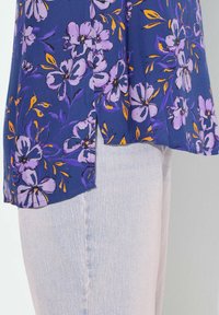 Blusa floreale con sfondo blu scuro e fiori viola e arancioni. Il tessuto è morbido e l'orlo presenta spacchi laterali per un tocco di dettaglio in più.