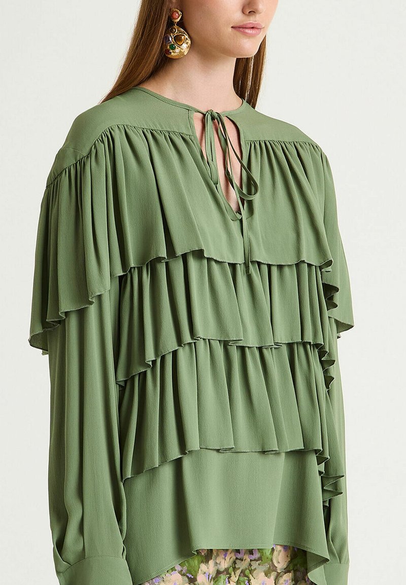 Grüne Bluse aus leichtem Stoff mit mehreren horizontalen Rüschen, einem Schlitz am Ausschnitt und Bindebändern, mit langen Ärmeln.