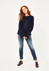 Dunkelblauer Strickpullover mit weitem Halsauschnitt und langen Ärmeln, kombiniert mit hellblauen Jeans, die an den Bündchen hochgerollt sind, und braunen Lederschuhen.