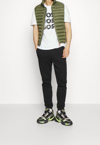 Chaleco acolchado verde oliva sobre una camiseta blanca con texto negro, combinado con pantalones deportivos negros y zapatillas negras con acentos en verde y gris.
