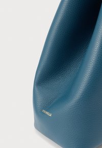 Υφασμένη μπλε δερμάτινη τσάντα με μικρό χρυσό λογότυπο "Coccinelle" εγχαραγμένο κοντά στην κάτω γωνία σε λευκό φόντο.