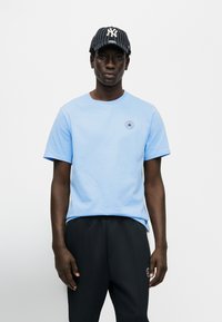 Ανοιχτό γαλάζιο βαμβακερό μπλουζάκι με στρογγυλή λαιμόκοψη και κοντά μανίκια, με ένα μικρό κόκκινο και μπλε αστέρι λογότυπο στο αριστερό στήθος. Μαύρο καπέλο με λευκές ρίγες.