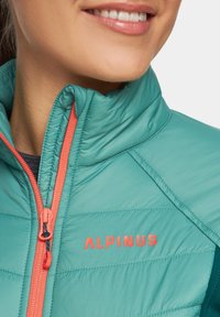 Turkusowa kamizelka insulationowa z wysokim kołnierzem, pomarańczowym zamkiem błyskawicznym i kontrastowym logo. Gładka faktura z przeszyciami i wyraźnymi szwami.