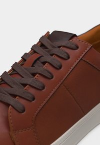 Καφέ δερμάτινο αθλητικό παπούτσι με σκούρα καφέ κορδόνια, υφασμένο πλαϊνό πάνελ και λευκή λαστιχένια σόλα, εμφανιζόμενο σε κοντινή λήψη από πάνω.