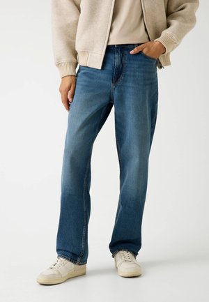 Jeans en denim bleu avec un design à jambe droite, présentant des coutures visibles, une taille classique et une texture usée. Associés à des baskets blanches.