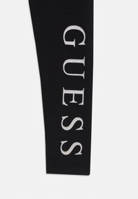 Schwarze Leggings mit einem glänzenden silbernen "GUESS"-Logo, das senkrecht an der Außenseite des linken Beins aufgedruckt ist. Entworfen für einen figurbetonten Look.