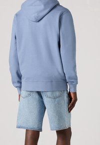 Felpa leggera blu con tasca a marsupio, polsini a costine e cappuccio con lacci, abbinata a pantaloni corti in denim chiaro. Texture liscia e vestibilità casual.