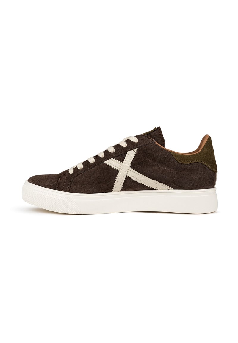 Sneaker in suede marrone con cuciture e lacci bianchi, caratterizzata da un design a croce prominente e un accentuato tallone verde a contrasto. Suola in gomma bianca.