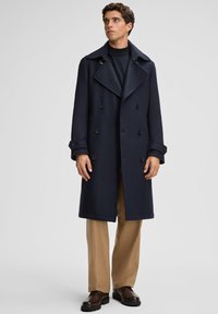 Man in een marineblauwe double-breasted jas, zwarte turtleneck, beige broek en donkerbruine leren schoenen staand op een effen achtergrond.