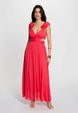 Donna in piedi che indossa un abito rosso fluente senza maniche, con dettagli arricciati sulle spalle e ritagli in vita, abbinato a sandali dorati e braccialetti.