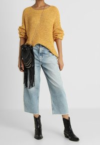 Chunky knit yellow sweater with rolled sleeves, light blue wide-leg jeans with frayed hem, black bucket bag with fringe, and glossy black boots.