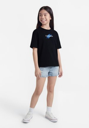 Fată zâmbitoare stă în picioare purtând un tricou negru cu imprimeu, pantaloni scurți din denim deschis, șosete albe și adidași albi, pe fundal simplu alb.