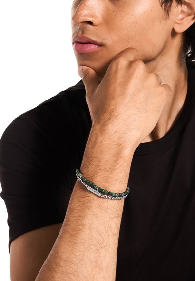 Bracelet de perles vertes superposé avec une chaîne de ton argenté. Comprend des pierres rondes et une étiquette en métal, porté au poignet contre une chemise noire.