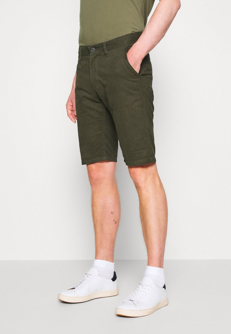 Shorts beige en tissu texturé, coupe droite, poches latérales et fermetures à bouton. Associés à des baskets blanches et des chaussettes de cheville.