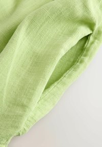 Tissu en lin vert clair avec une surface texturée, présentant de doux plis et un léger éclat. Le bord est cousu avec une finition soignée.