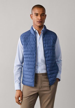 Man wearing light blue button-up shirt, blue quilted vest, and brown pants, standing against plain light gray background.