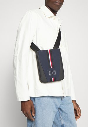 Sac bandoulière bleu marine avec un design texturé, comprenant une bande verticale d'accent rouge et blanche, et une sangle noire. Base noire unie, détail du logo.