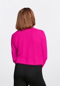 Cárdigan de punto fucsia con un diseño corto y dobladillo acanalado, con mangas raglán. Se lleva con pantalones negros ajustados. Vista trasera.
