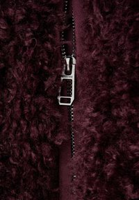 Burgunderfarbenes, zotteliges Stoffmaterial mit einer weichen, strukturierten Oberfläche; verfügt über einen Metallreißverschluss mit rechteckigem Zuggriff in der Mitte.