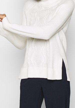 Personne portant un pull à col roulé blanc à torsades et un pantalon foncé, tendant un bras pour ajuster la manche.