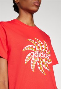 Röd bomullströja med geometrisk tryck i gult, rosa och vitt, med "adidas"-logotypen centrerad i designen. Kortärmad.