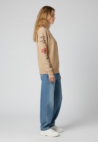 Felpa beige con testo e disegni di cuori rossi sulle maniche, abbinata a jeans blu larghi e sneakers bianche. Texture liscia.