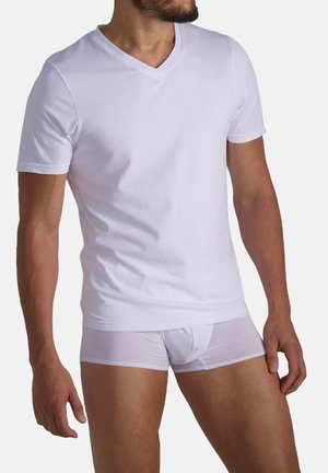 Mann in einem taillierten weißen V-Ausschnitt-T-Shirt und weißen Boxer-Shorts, der vor einem schlichten hellen Hintergrund steht.