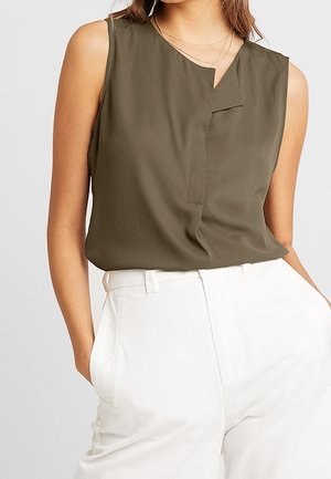Blouse sans manches vert olive avec un plissé subtil à l'avant, associée à un pantalon blanc taille haute, mains dans les poches.