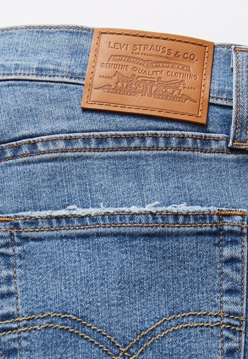 Jean en denim bleu clair avec un patch en cuir marron. Le patch présente un texte en relief et des détails de design. Couture dorée tout au long.