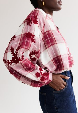 Rood-wit geruit overhemd met lange, opgeblazen mouwen met textuur van rode bloemrijke borduursels langs de schouders en manchetten. Casual pasvorm, gecombineerd met donkere denim.