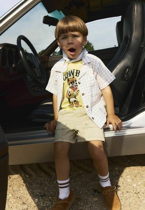 Kind gekleed in een gele graphic t-shirt, een licht geruit overhemd met korte mouwen, beige shorts, witte sokken met bruine strepen en bruine schoenen, zittend in een auto.