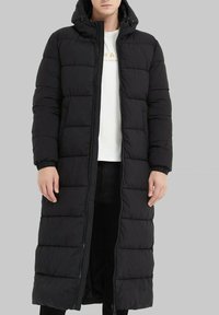 Manteau long noir de type doudoune avec capuche, comportant des sections matelassées, des poignets élastiques et une fermeture éclair à l'avant, porté sur une chemise blanche et un pantalon noir.