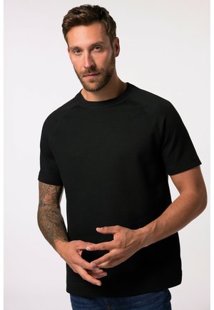 T-shirt noir à manches courtes avec col rond et manches raglan, fabriqué en tissu texturé. Le modèle pose les mains ensemble, mettant en valeur la coupe.