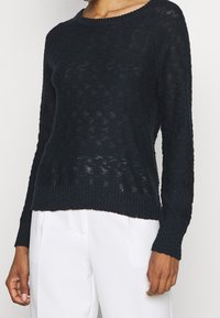 Tmavě námořnický pletený svetr s volným, texturovaným vzorem, kulatým výstřihem a žebrovanými manžetami a lemem, kombinovaný se světle zbarvenými kalhotami.