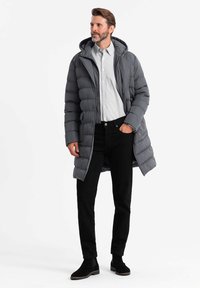 Cappotto imbottito grigio con cappuccio, caratterizzato da un design trapuntato e chiusura con zip. Indossato sopra una maglietta grigia chiara e pantaloni neri, con stivali neri.