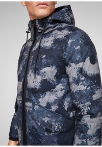 Vadderad jacka i marinblå och grå camouflage-mönster, med dragkedja framtill, huva och sidofickor med tryckknappslås. Slät tygtextur.