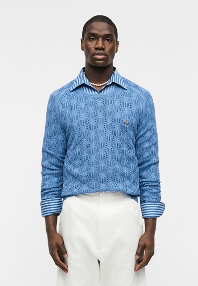 Homme portant un pull en maille à motifs bleu clair sur une chemise à col rayée bleue, un pantalon blanc et un collier de perles sur un fond uni.