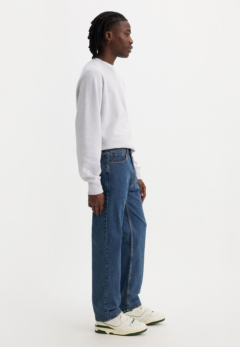 Un sweat-shirt gris à col rond, un jean droit bleu et des baskets blanches avec des accents verts. La vue de côté montre une coupe décontractée et un style casual.