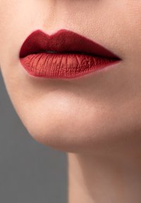 Labios carnosos pintados con un lápiz labial rojo mate profundo, con un arco de cupido bien definido y una ligera textura, sobre un fondo de tono de piel neutro y suave.