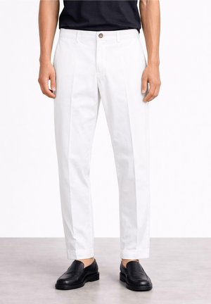 Mężczyzna w białych, dopasowanych spodniach, czarnych mokasynach i czarnej koszuli włożonej do środka, stojący na jasnoszarej podłodze przed białą ścianą.