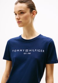 Tamsiai mėlynos spalvos medvilninis marškinėliai su baltu spausdintu logotipu "TOMMY HILFIGER EST. 1985" krūtinės srityje ir mažuoju logotipo akcentu ant rankovės.